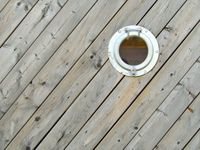 An orthogonal view of a wall made of diagonal wooden boards interrupted by a single porthole in the upper-right quadrant of the composition.