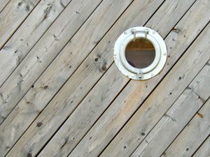 An orthogonal view of a wall made of diagonal wooden boards interrupted by a single porthole in the upper-right quadrant of the composition.