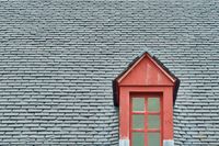 A window framed in red wood sticks out alone surrounded by dark grey slate roof tiles which make up most of the minimalit composition.