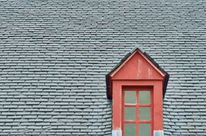 A window framed in red wood sticks out alone surrounded by dark grey slate roof tiles which make up most of the minimalit composition.