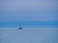 Many shades of blue from the sky and water meet in the middle of the photo where a small sailboat sits on the horizon.