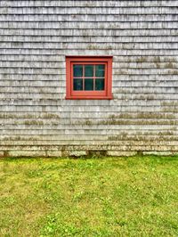 An orthogonal view of a small red window in the middle of a grey, weathered wall.