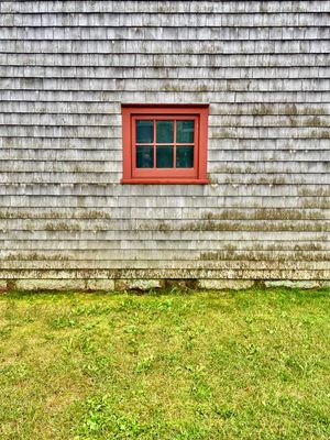 An orthogonal view of a small red window in the middle of a grey, weathered wall.