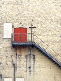 An orthogonal view of bright red double doors very high up on a brick wall with a black fire escape leading down.