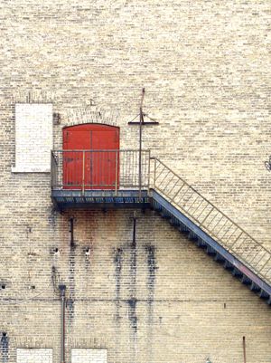 An orthogonal view of bright red double doors very high up on a brick wall with a black fire escape leading down.