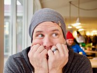 Head and shoulders portrait of a man in a hoodie and winter hat. His chin is resting in his hands and his fingers are covering his mouth, while his eyes look playfully to the side.