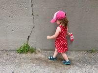 A small child is walking along a cracked concrete wall. She's wearing a sundress covered in Canadian mape leaf symbols and she's dragging a Canada flag along the wall.