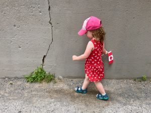 A small child is walking along a cracked concrete wall. She's wearing a sundress covered in Canadian mape leaf symbols and she's dragging a Canada flag along the wall.