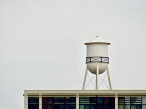 A rooftop with a white cylindrical water tower standing alone against a white overcast sky.