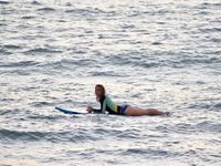 In between waves a woman looks into the camera while laying on her stomach on a surfboard.