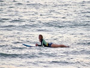 In between waves a woman looks into the camera while laying on her stomach on a surfboard.