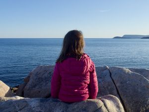 A young girl sits on a rock with her back to the camera as she looks at the Atlantic Ocean.