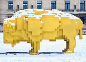 Side view of an outdoor art installation of a bright yellow abstract bison made of cunic blocks. Snow rests on top of the sculpture.