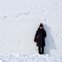 A woman dressed in black winter outerwear stands against a flat wall of white snow that's twice her height. The wall is pockmarked with dents and cracks in the snow.