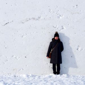 A woman dressed in black winter outerwear stands against a flat wall of white snow that's twice her height. The wall is pockmarked with dents and cracks in the snow.
