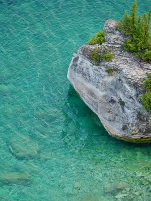 Looking down at a granite boulder sitting in crystal clear blue-green water. The tropical look of the lake contrasts with the pine trees perched atop the rock.