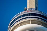Looking up at the CN Tower from the ground. Some thrill-seeking tourists can be seen in orange jumpsuits walking around the edge against a lush blue sky.