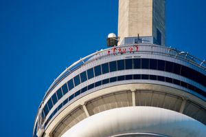 Looking up at the CN Tower from the ground. Some thrill-seeking tourists can be seen in orange jumpsuits walking around the edge against a lush blue sky.