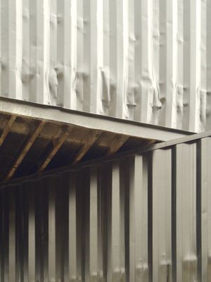 Looking up at 2 black shipping containers. The upper container is at an angle to the lower one and overhangs such that the underside forms a triangular shape in the center of the frame.