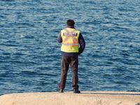 A security guard's back (and bright saftey vest) is to the camera as he watches the ocean which fills the entire background of the photo.