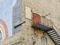 An angled view of bright red double doors very high up on a brick wall with a black fire escape.