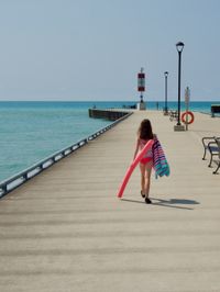 A child walks away from the camera towards the end of a lakeside pier. She carries herself with determination and she's holding a pool noodle and beach towel.