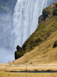 The view of a tall waterfall is obstructed by a rocky hillside covered in moss and grass in the foreground.