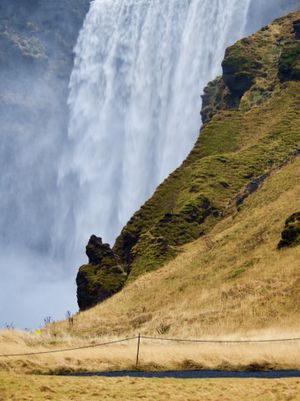 The view of a tall waterfall is obstructed by a rocky hillside covered in moss and grass in the foreground.