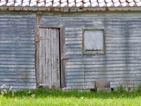 An orthogonal view of an old, grey shack with a boarded up window and a makeshift door attached with rusty hinges.