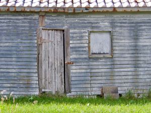 An orthogonal view of an old, grey shack with a boarded up window and a makeshift door attached with rusty hinges.