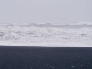 Looking at the ocean on a very foggy day. The photo is framed in three equal segments—the sky is light grey, the water is grey and white, and the ground is black volcanic sand.