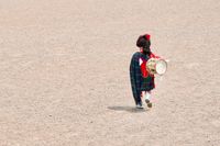 A candid shot looking down at a Scottish marching band drummer walking across a gravel parade ground. He's wearing full Highland dress and a kilt, and carrying a drum over his shoulder.