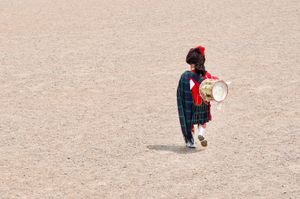 A candid shot looking down at a Scottish marching band drummer walking across a gravel parade ground. He's wearing full Highland dress and a kilt, and carrying a drum over his shoulder.
