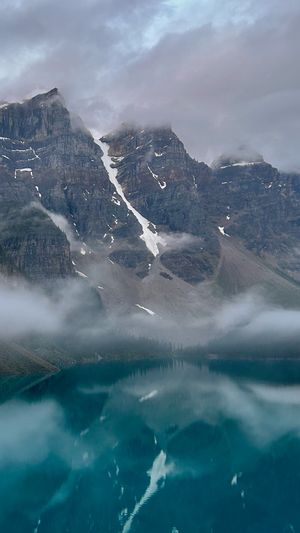 The snowy mountain peaks stand tall above Moraine Lake's crystal blue water as numerous cloud banks brush past them.
