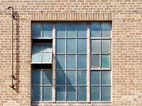 An orthogonal view of the brick wall of an industrail building. In the middle of the wall there's a large set of dirty windows and only one of them is tilted open.