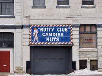 An orthogonal view of a beige warehouse building. A window at street-level is boarded up with an orange sign reading 'PANCAKE SYRUP'. In center frame is a loading door under a blue sign reading 'Nutty club' and featuring a character dressed in pinstripes and a red top hat.