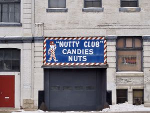 An orthogonal view of a beige warehouse building. A window at street-level is boarded up with an orange sign reading 'PANCAKE SYRUP'. In center frame is a loading door under a blue sign reading 'Nutty club' and featuring a character dressed in pinstripes and a red top hat.
