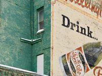 Angled view looking up at a large brick wall above an alley with a old, faded mural featuring a vintage advertisement for Pepsi-Cola.