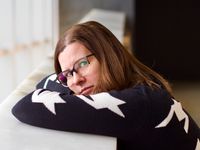 A head-and-shoulders portrait of a beautiful woman wearing glasses and resting her head on her crossed arms. She's staring pensively at the camera.