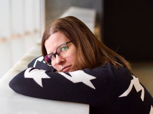 A head-and-shoulders portrait of a beautiful woman wearing glasses and resting her head on her crossed arms. She's staring pensively at the camera.