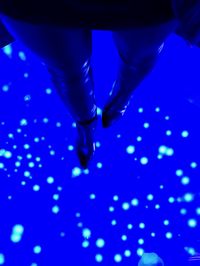 Looking down at a glowing blue glass floor with jellyfish floating beneath. Standing on the floor is a woman in black leather tights and boots, with only her legs in the frame.