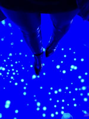 Looking down at a glowing blue glass floor with jellyfish floating beneath. Standing on the floor is a woman in black leather tights and boots, with only her legs in the frame.
