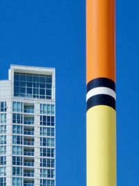 The top of a large fishing bobber in an outdoor art installation forms orange, black, white, and yellow stripes against a bright and sunny blue sky. A high-rise glass condo tower can be seen in the background.