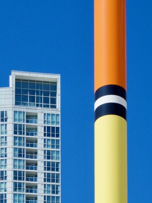The top of a large fishing bobber in an outdoor art installation forms orange, black, white, and yellow stripes against a bright and sunny blue sky. A high-rise glass condo tower can be seen in the background.