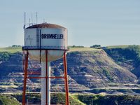 Landscape photo of the wall of a valley in the backround. IN the foreground is a white and red water tower that says 'DRUMHELLER' on the side of the reservoir.