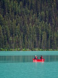 Two people paddle a red canoe across a glacial lake against a towering backdrop of tall evergreen trees.