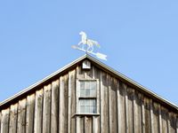 Looking up at a white weather vane shaped like a horse on top of an old barn.