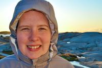 Portrait of a cute woman with her hood drawn tight around her face. The sun is rising behind her over the rocks of the rugged Nova Scotia coastline.