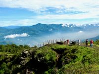 Landscape of a lush green mountain range covered in clouds, with some tourists walking a trail in the foreground.