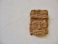 Close-up of a small Mayan stone carving of a face, exhibited by itself on a white plaster museum wall.